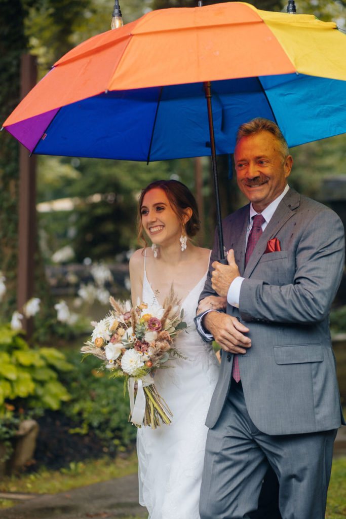 Person in suit walking the celebrant down the aisle holding a rainbow umbrella 
