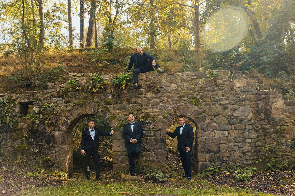groomsmen standing in front of stone wall with the groom sitting on top of it