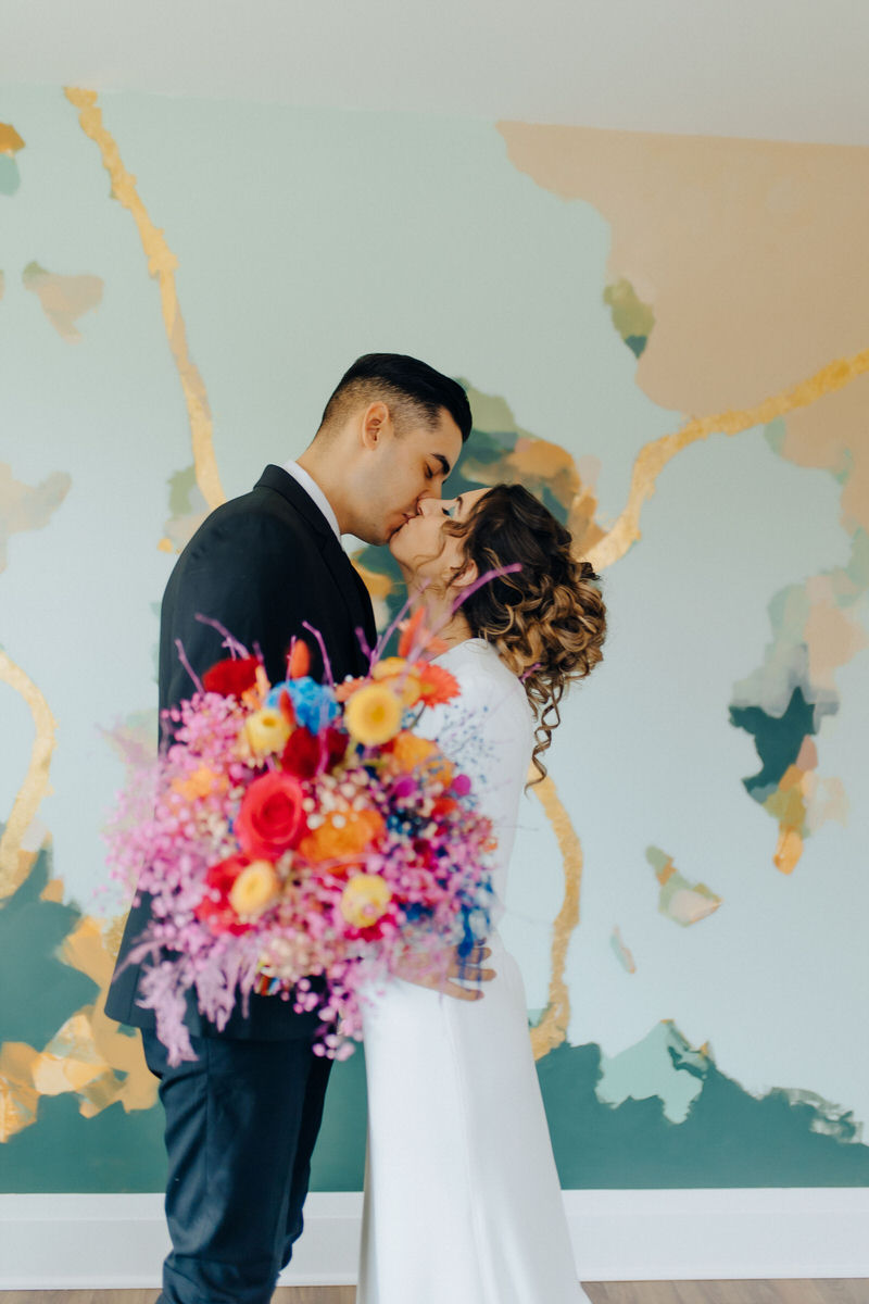 couple kissing while holding brightly colored flower bouquet 