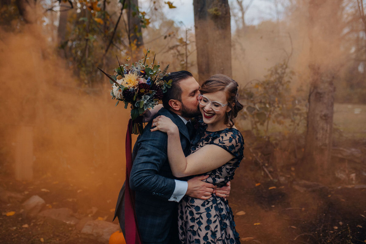 person kissing spouse on the cheek with orange smoke behind them