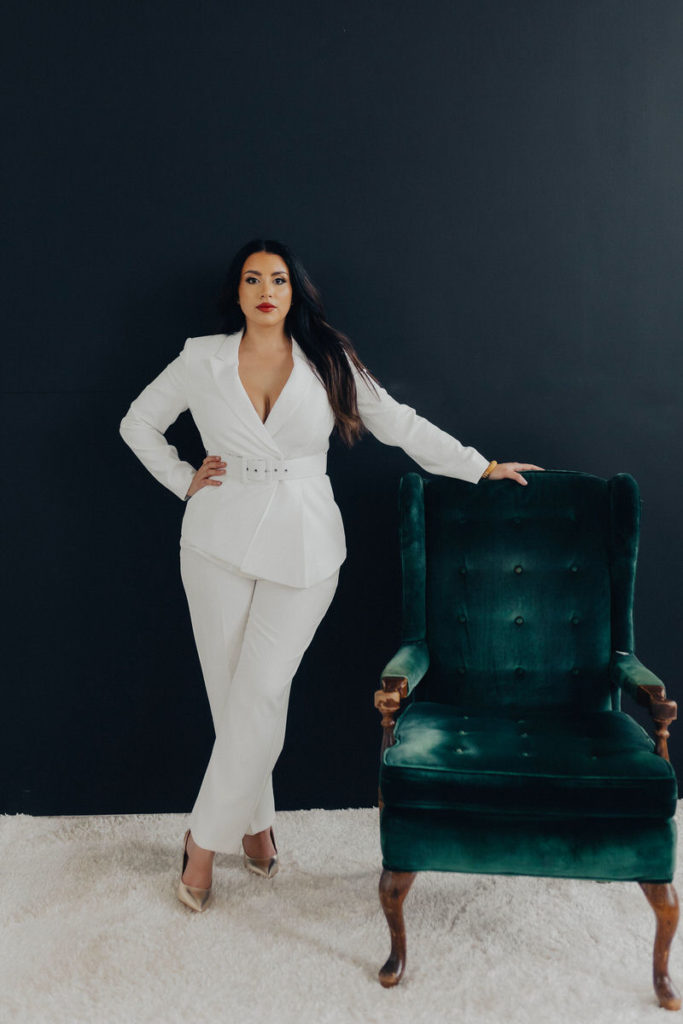 woman in an all white suit with her arm rested on the top of a green velvet chair