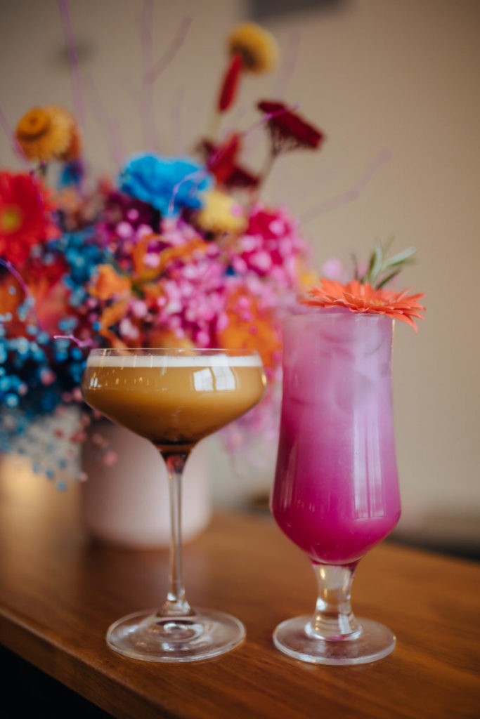 dark brown cocktail and bright pink cocktail sitting in front of a colorful bouquet of flowers 