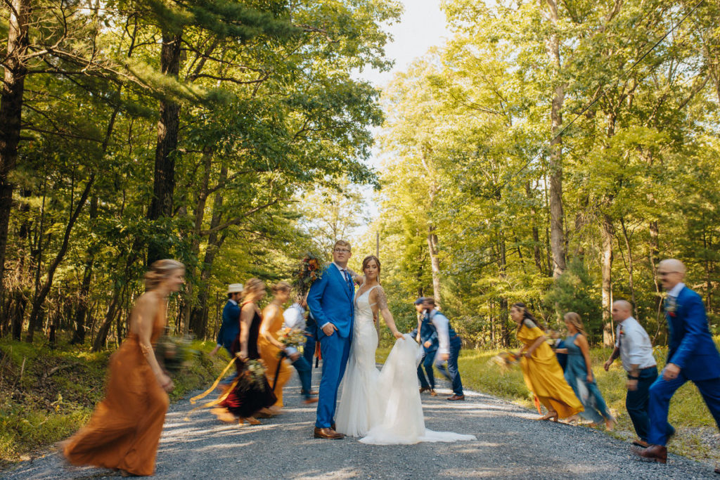 wedding couple standing in a middle of a forest road while their wedding party runs on either side of them while slightly blurred 