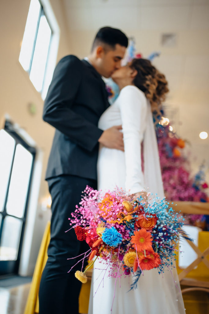 bright blue, orange, and pink bouquet behind held by a person who is kissing their partner 