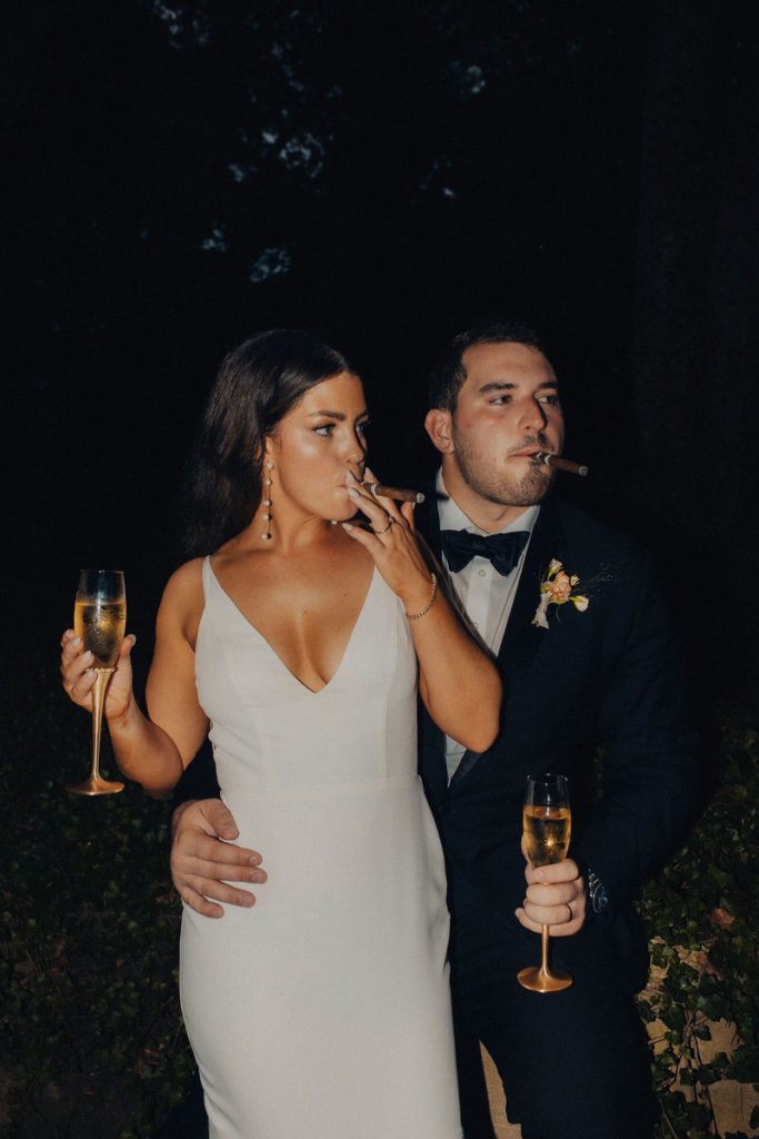 person with their arm around their spouse's waist as they both hold a glass of champagne and smoke a cigar 