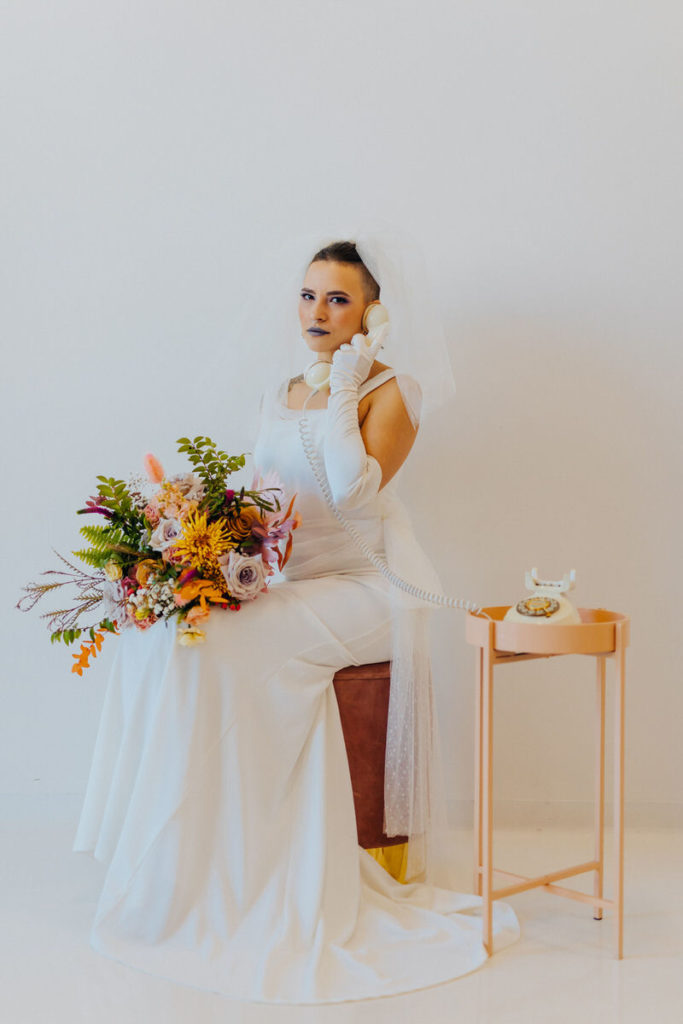 person in a wedding dress sitting on a stool while holding a bouquet with one hand and a phone to their ear in the other