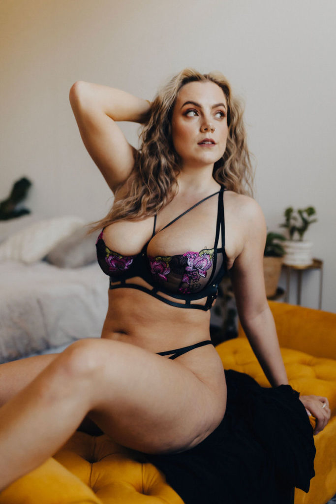 person sitting on a couch in lingerie leaning back on one hand and holding the other up to their head 