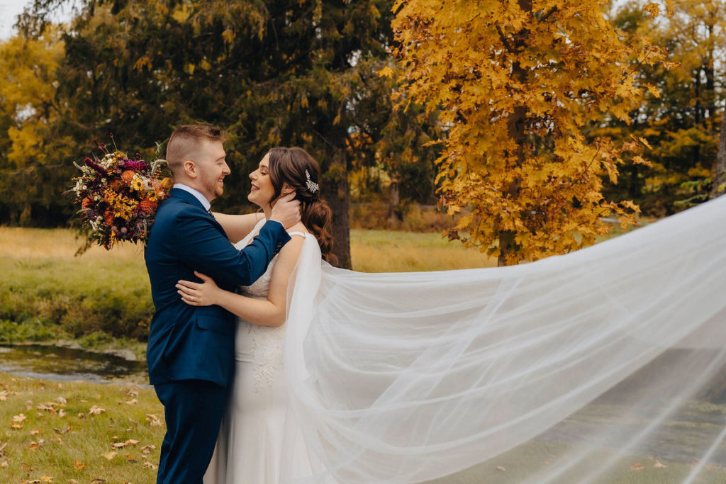 couple with their arms around each other about to kiss as the veil of their dress flows behind them in the wind 