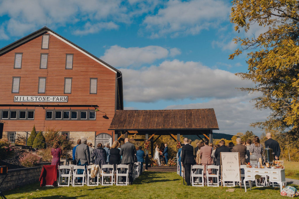 wedding guests standing up as they look towards the couple standing up at the alter in front of a large red farm building 