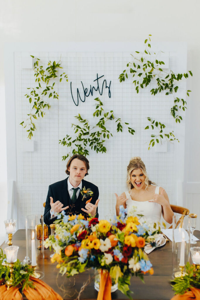 A married couple sitting at the end of a reception table while holding up shaka signs. 