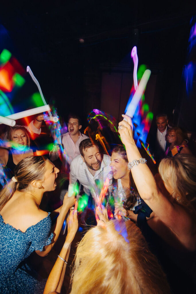 A group of people at a wedding dancing, singing, and holding up light sticks. 