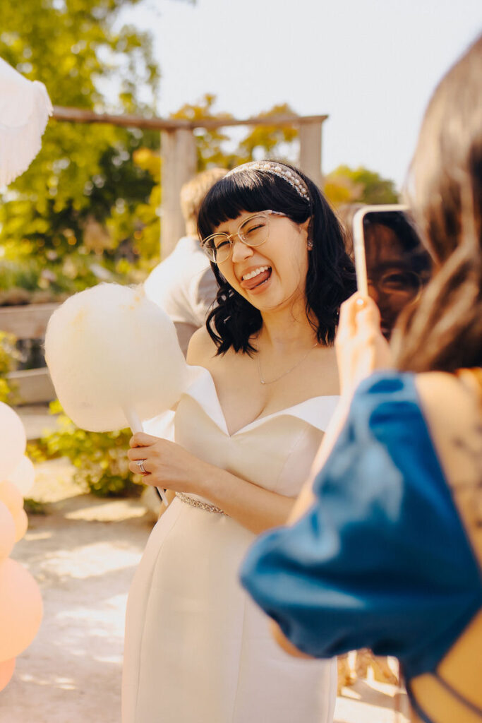 A person in a wedding dress smiling and holding cotton candy as someone takes their picture on a phone. 