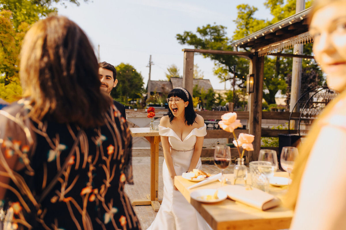 Candid wedding photography of a bride laughing as guests stand around them while outside.