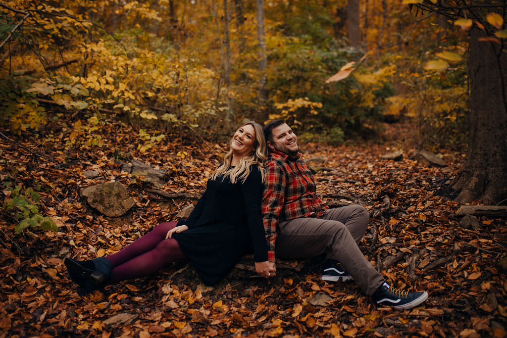 A couple sitting back to back on a leaf covered ground. 