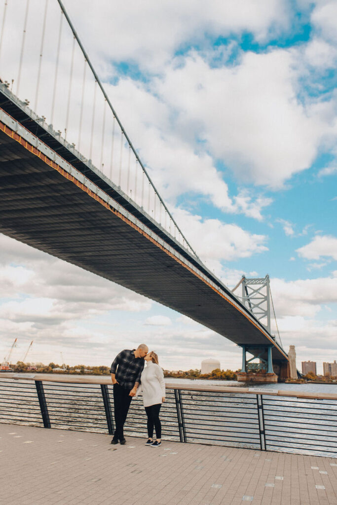 A couple kissing with a large bridge behind them. 