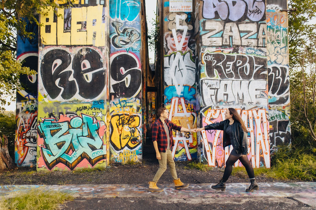 A couple holding hands and walking along a path next to a bridge covered in graffiti. 
