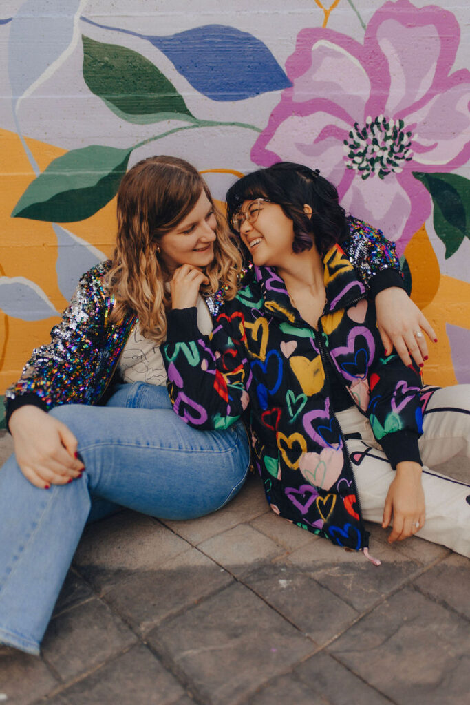 A couple sitting on the ground in front of a colorful mural leaning in for a kiss. 