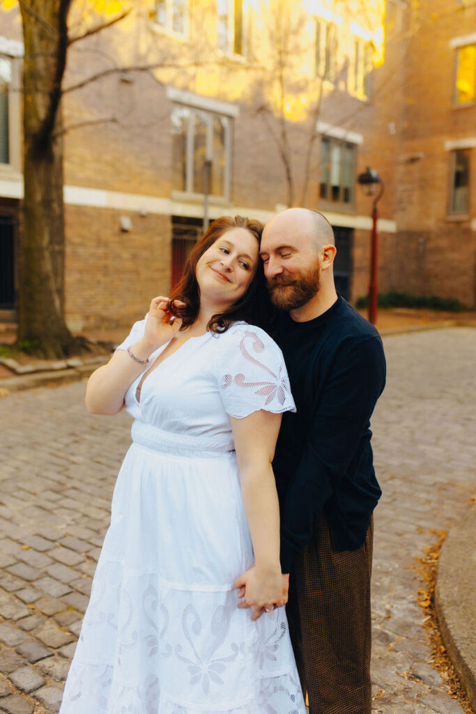 A couple smiling while holding hands and standing on a cobblestone street. 