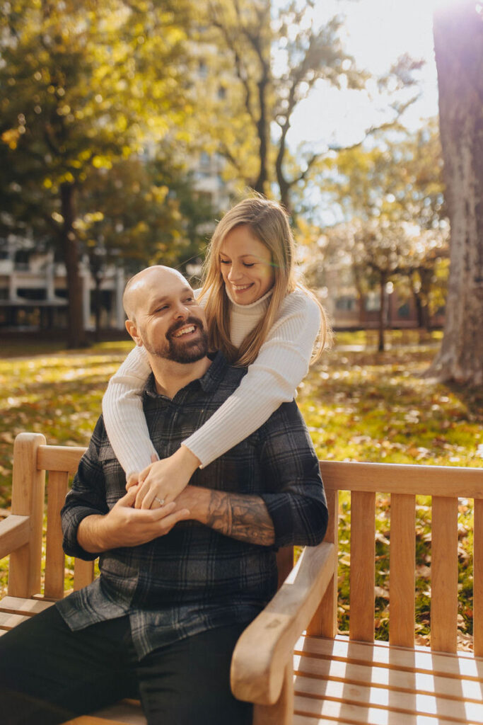 A person sitting on a bench with their partner standing behind them with their arms over their shoulders. 