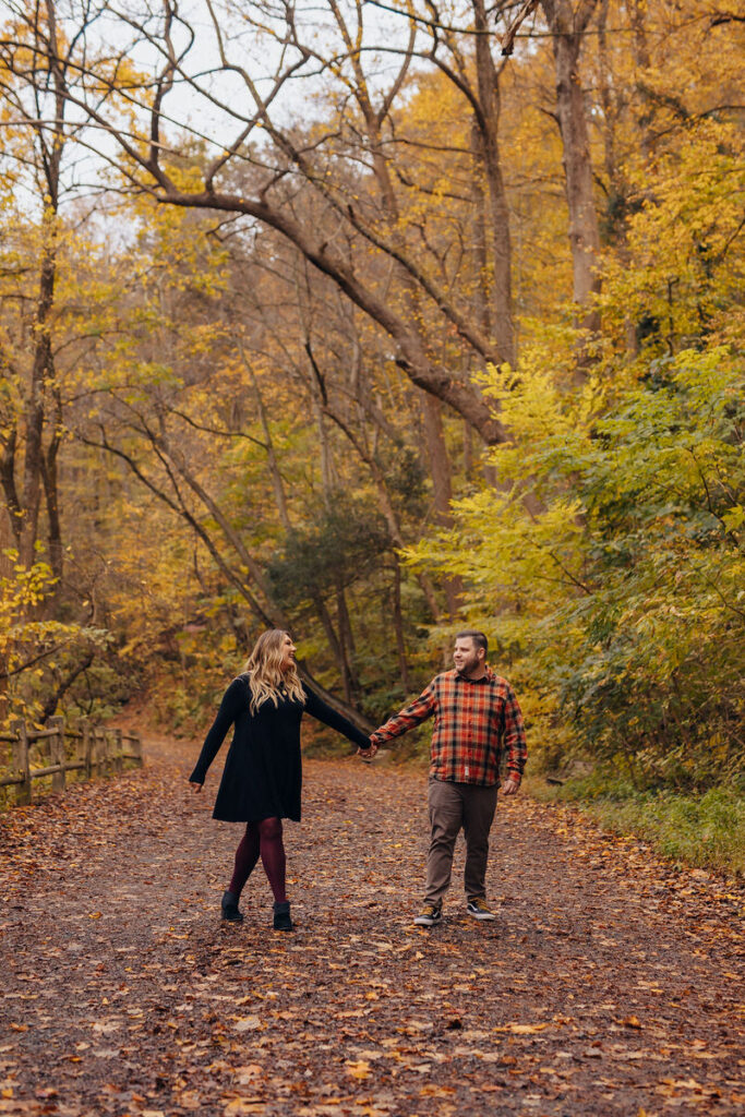 A couple holding hands and walking along a leaf covered path. 