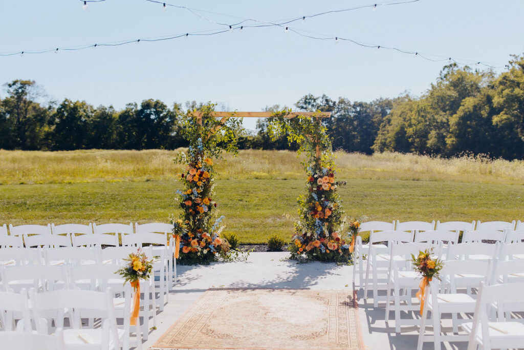 A flower covered arch at the end of an aisle of chairs in an open field wedding venue. 