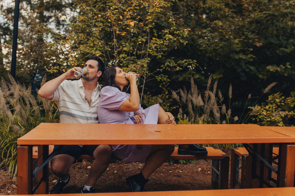 A couple sitting at a picnic table drinking out of cans.
