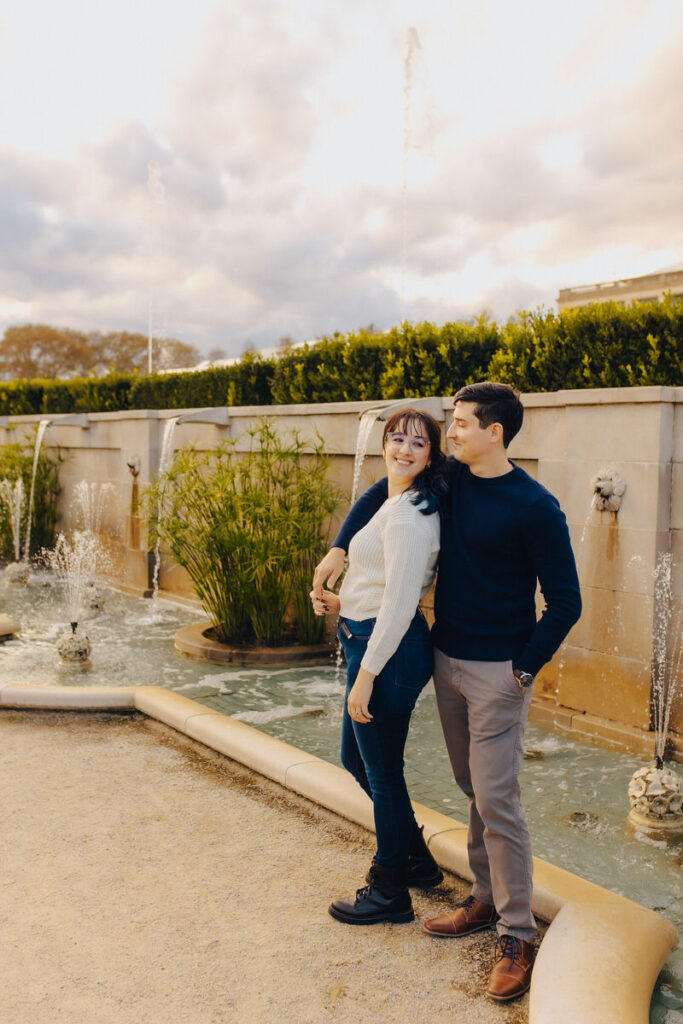 A person with their arm around their partner while they stand next to a fountain. 