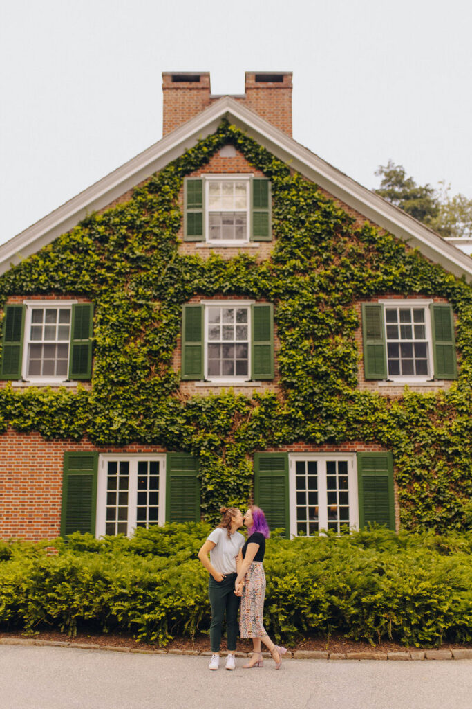 A couple about to kiss while standing in front of a vine covered house.