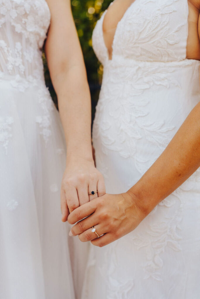 Detail of two people holding hands, featuring their wedding bands and the intricate lace patterns of their gowns.
