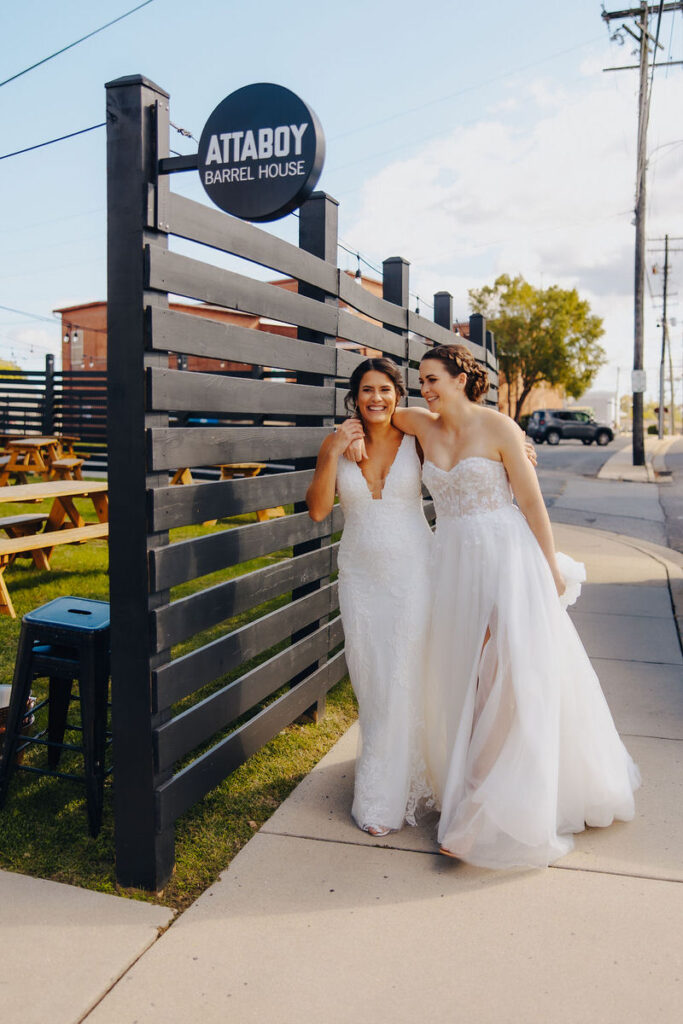 A wedding copule laughing and walking past a brewery sign, sharing a joyous moment in their wedding dresses.
