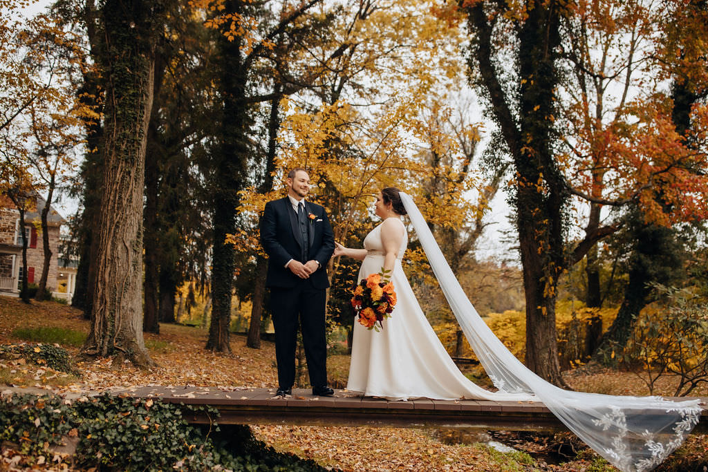 A wedding couple standing apart on a leaf-covered bridge in a fall setting, gazing into each other's eyes, with a picturesque house in the background.
