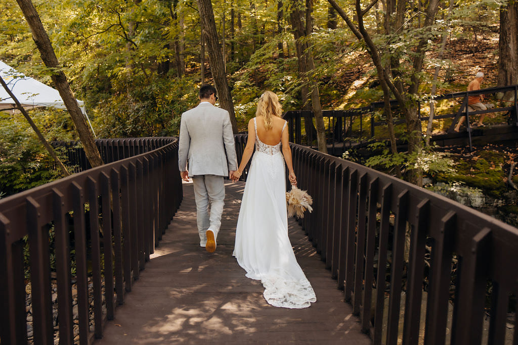 A couple walks hand in hand along a bridge surrounded by lush trees, with one person wearing a long white gown holding a bouquet, and the other in a light grey suit.
