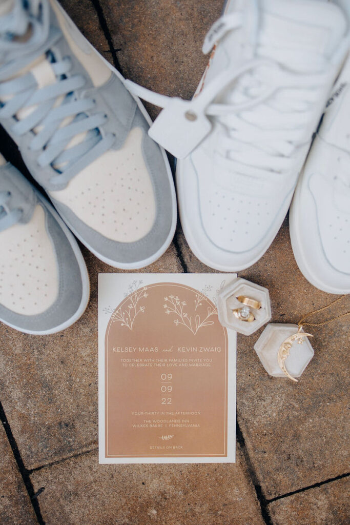 Wedding shoes beside an invitation card, with the names "Kelsey Maas & Kevin Zwaig" and a date, accompanied by jewelry boxes and a necklace.
