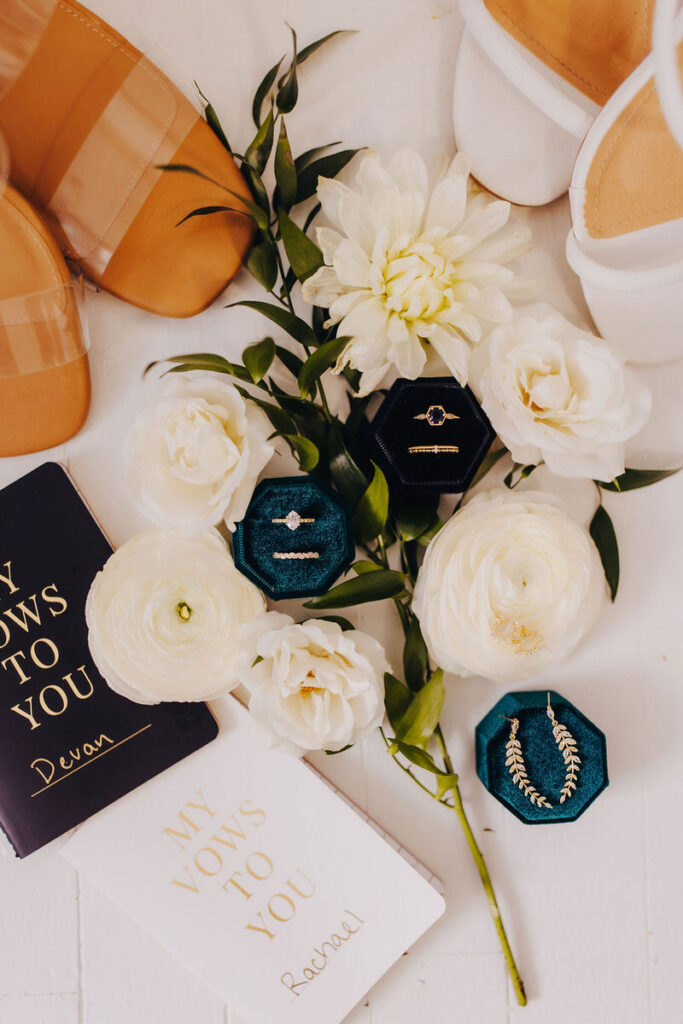 A flat lay arrangement of wedding vow books titled "My Vows to You," with a pair of transparent heels, white heels, white flowers, and two ring boxes.
