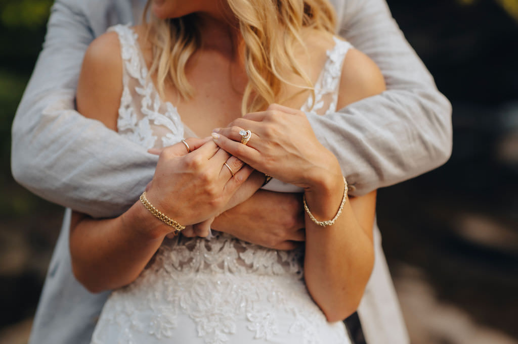Close-up of a couple's hands entwined, showcasing their wedding rings against a lace gown.