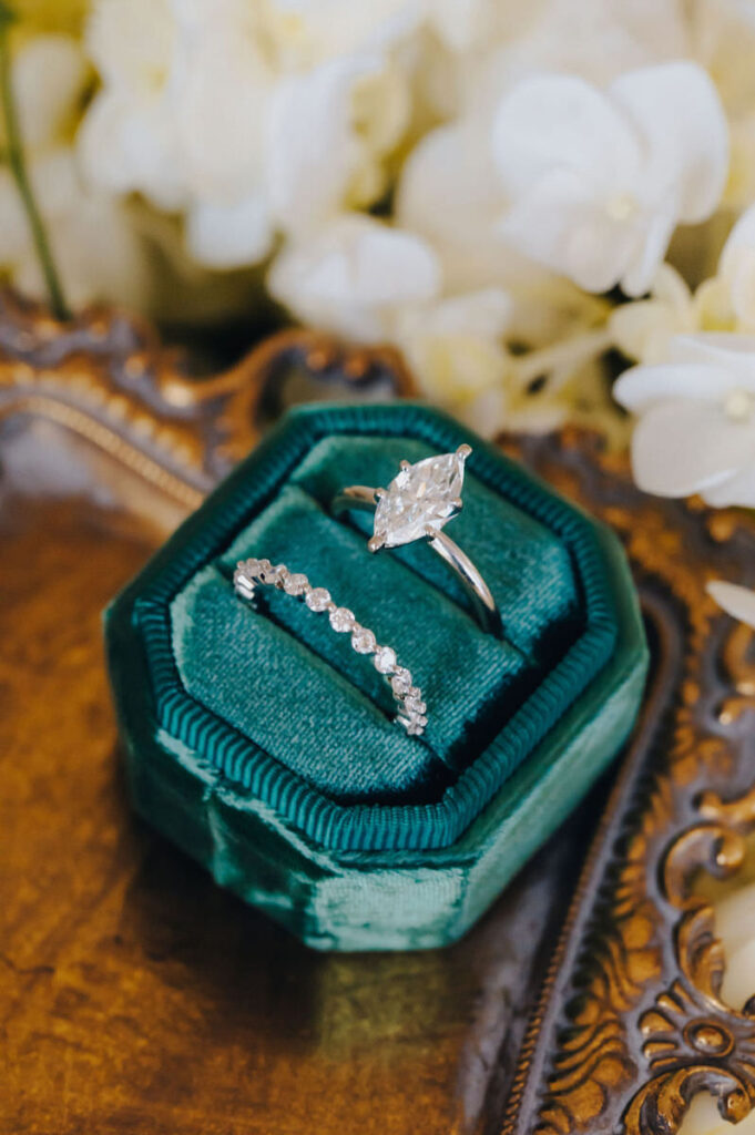 Marquise diamond engagement ring and wedding band displayed in a green velvet box with white orchids in the background.