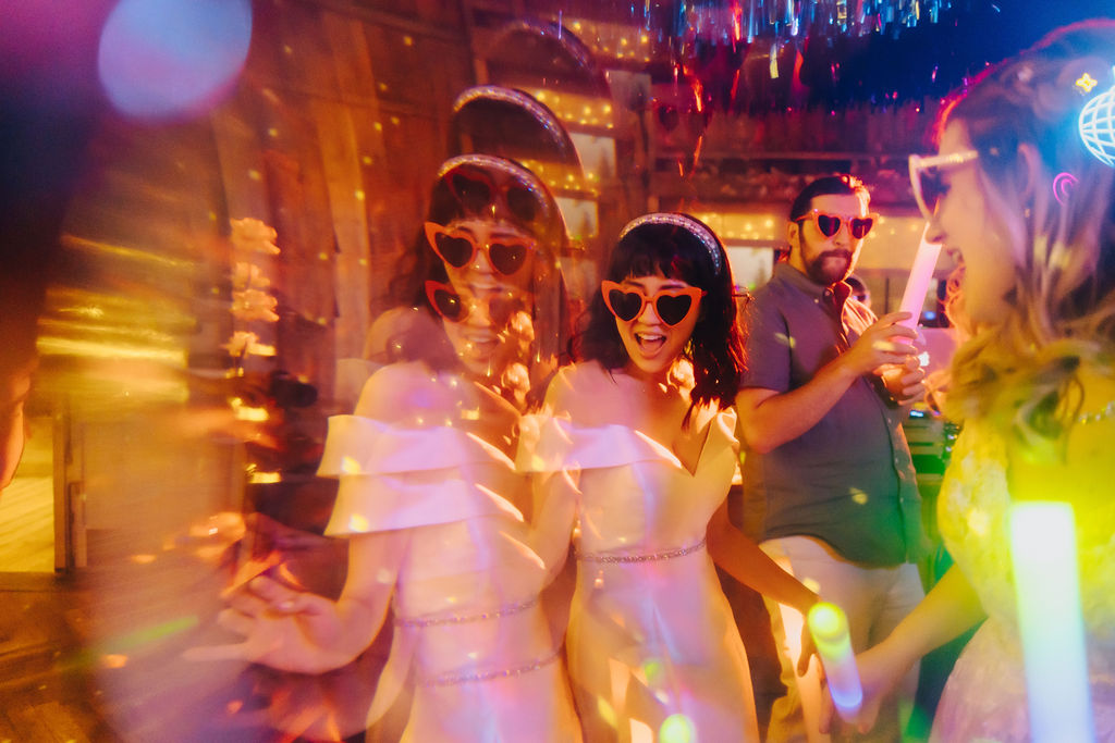 Wedding guests with heart-shaped sunglasses dancing under warm lights at a vibrant party.