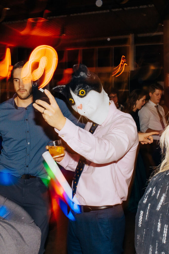 Party attendee in a whimsical cat mask taking a selfie, with light trails and fellow partygoers in the background.

