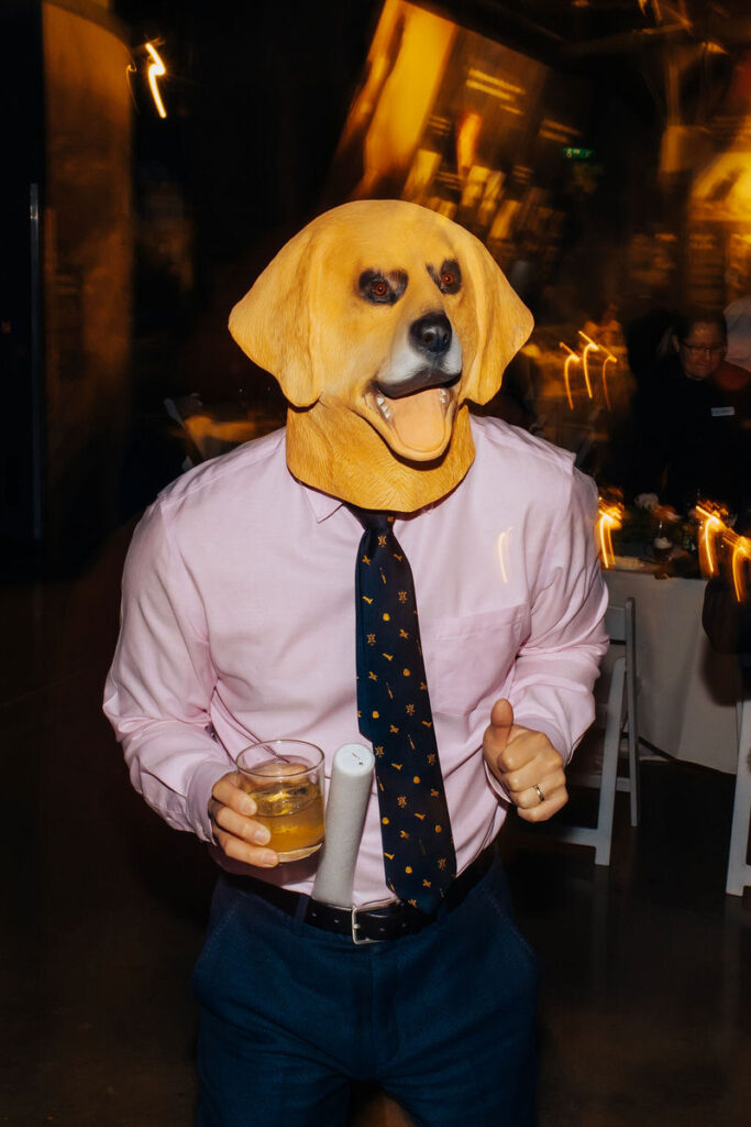 Person wearing a realistic dog mask holding a beverage, surrounded by the ambient lighting of a social event.
