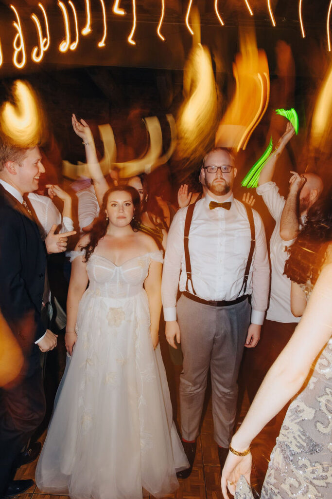 A wedding couple standing in the middle of the dance floor as their guests dance around them. 
