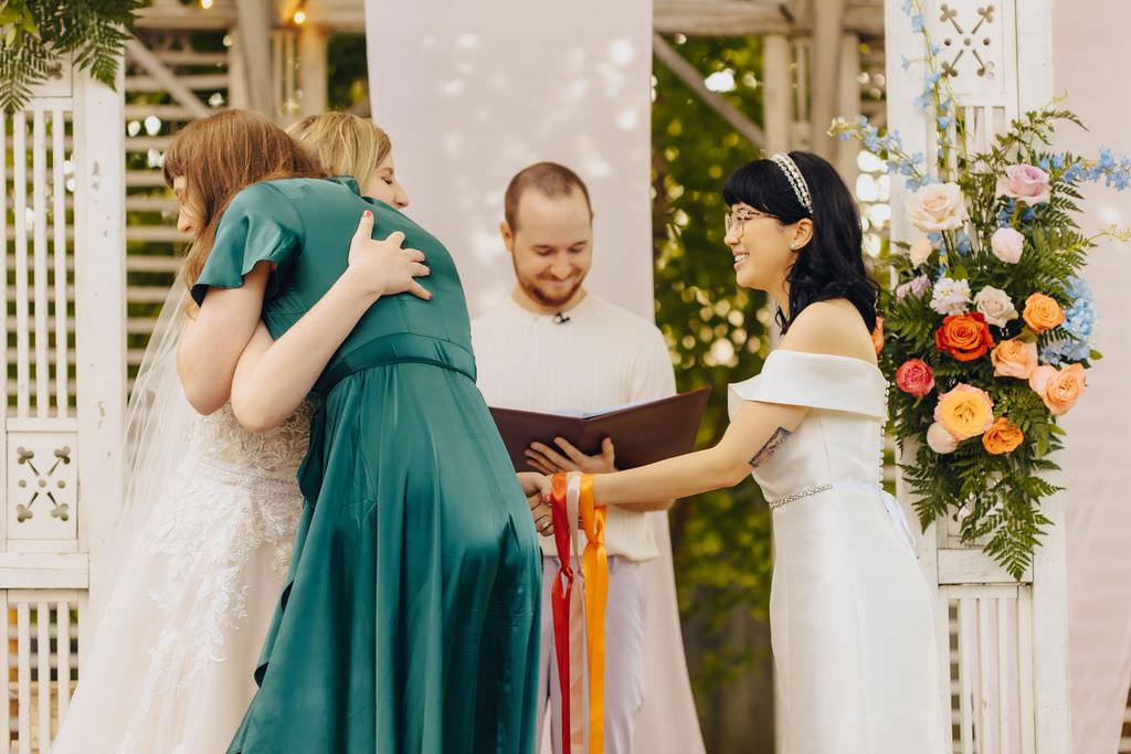 A heartfelt moment as a person in a teal dress hugs another, while another person and the officiant share a joyful look during an outdoor wedding ceremony.