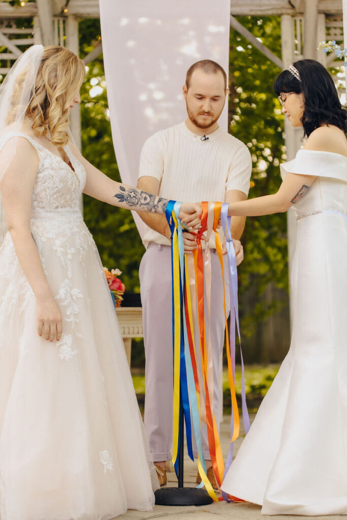 Two partners and an officiant holding a handfasting ceremony with colorful ribbons, symbolizing unity, in an outdoor wedding setting.