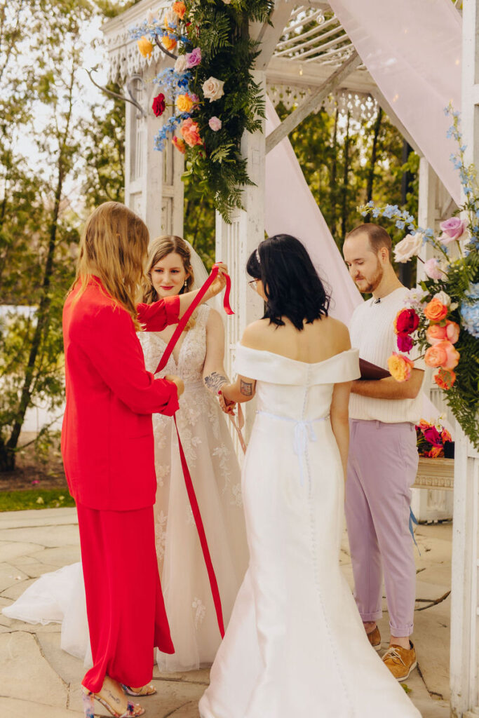 A to-be-wed in a lace gown receiving a red ribbon from another to-be-wed in a red dress, part of a wedding handfasting ritual, with an officiant and another person observing.