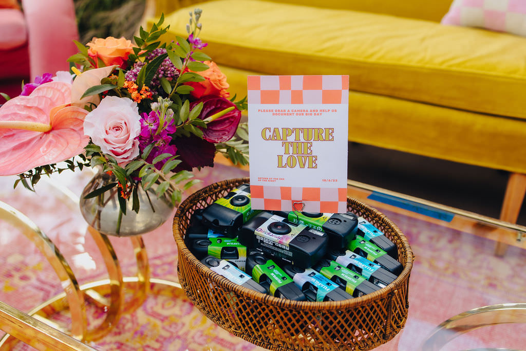 A colorful wedding setup featuring a bouquet of flowers in a vase, disposable cameras in a wicker basket, and a 'Capture the Love' sign encouraging guests to take photos, with a plush yellow couch in the background.
