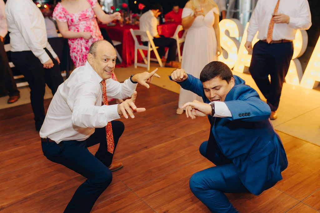 Two people in high spirits, one in a white shirt and festive tie and the other in a blue suit, playfully pose with dance moves on the dance floor at a wedding reception.