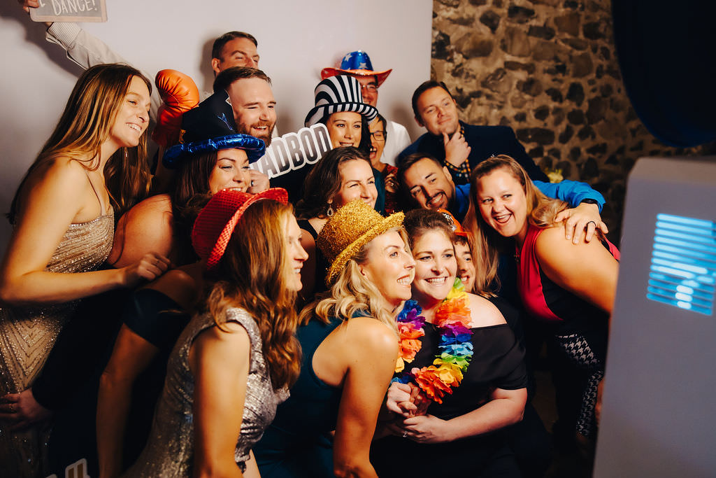A lively group of wedding guests wearing playful hats and props, huddled together for a fun photo at a photo booth, exuding joy and celebration.