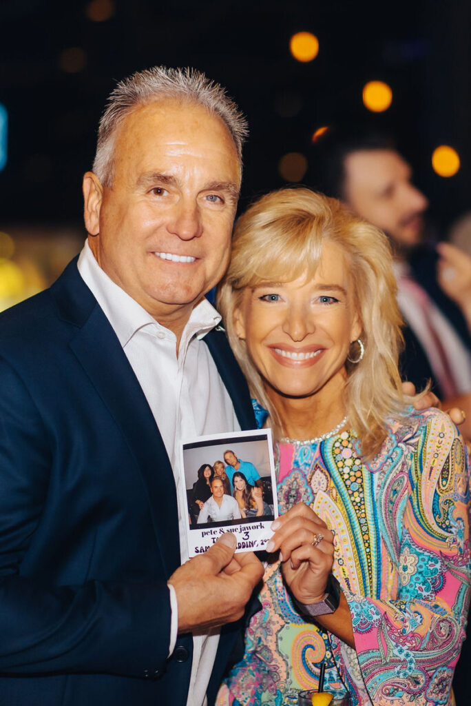 A smiling couple holding a Polaroid photo of themselves at a wedding reception, with a backdrop of guests and warm ambient lighting.