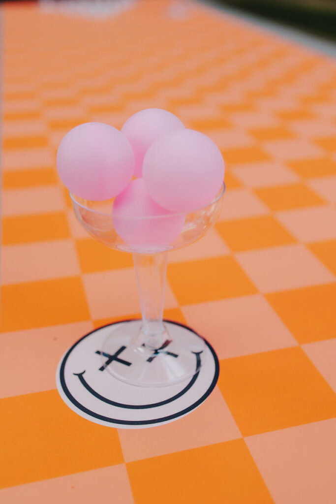 A close-up of a transparent wine glass on an orange and white checkered table, holding three pink balls, part of a wedding table game setup.