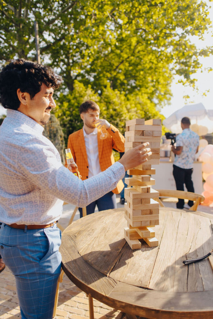 A person in a patterned shirt and blue trousers carefully playing a giant game of Jenga at an outdoor wedding reception, with a guest in the background sipping a drink.