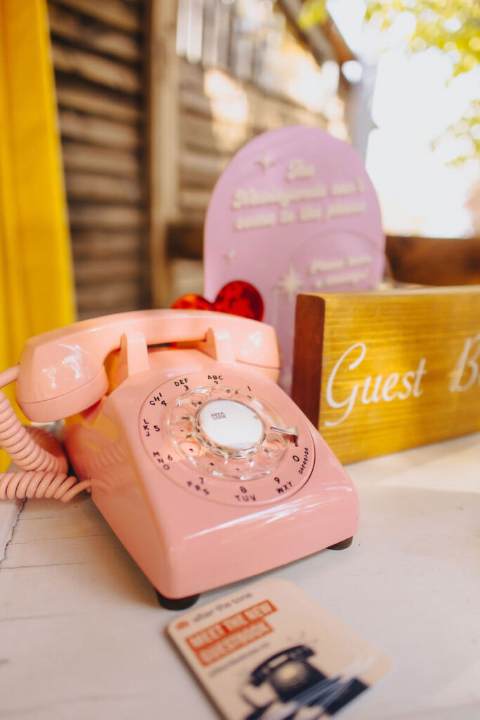 A vintage pink rotary phone set against a blurred backdrop featuring a heart-shaped sign and 'Guest Book' in cursive writing, suggesting a whimsical wedding guest interaction area.